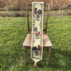 May include: A decorative table runner featuring a repeating pattern of colorful roosters and floral accents on a light yellow background. The runner is draped over a wooden chair and has a tassel at the bottom.