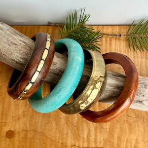 May include: Four wooden bangles in varying colors and designs. One is dark brown with gold-colored metal accents, another is turquoise, a third is hammered gold-colored metal, and the last is a glossy brown. The bangles are displayed on a piece of driftwood.