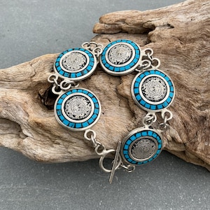 Peut inclure: Un bracelet à maillons argentés composé de cinq médaillons circulaires. Chaque médaillon présente un motif de calendrier en relief détaillé, entouré d'une bordure de petites incrustations de pierre rectangulaires bleu vif. Fermoir à bascule.