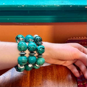 May include: A turquoise beaded bracelet with silver accents. The bracelet features large, round beads with a speckled pattern. The beads are strung together with silver-colored metal. The bracelet is worn on a wrist.