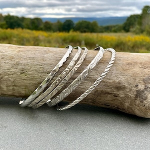May include: A set of four silver-toned metal bangles featuring distinct textures. The collection includes a faceted geometric band, a band with an intricate floral pattern, a band with a repeating leaf motif, and a twisted rope-style metal band.