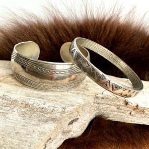Puede incluir: Dos pulseras de plata. Una es un brazalete ancho con líneas horizontales grabadas. La otra es una pulsera fina y abierta con un patrón de triángulos repetidos. Ambas se exhiben sobre un trozo de madera flotante contra un fondo de piel marrón.