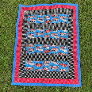 May include: A handmade quilt with a blue, red, and black color scheme. The quilt features a black background with white polka dots, red borders, and blue edges. Rectangular sections with a blue background display red and white fire trucks.