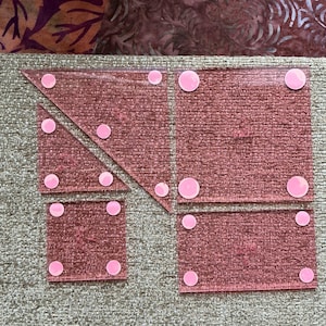 May include: A collection of pink quilting templates in various shapes, including squares and triangles. Each template has pink, circular, non-slip grips on the corners. The templates are arranged on a textured, neutral-colored surface.