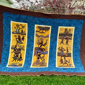 May include: A large, handmade quilt with a blue background and brown border. Three vertical panels feature yellow backgrounds with detailed illustrations of people, trees, and animals in black and brown. The quilt is displayed outdoors on a grassy surface.