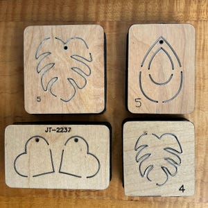 Könnte beinhalten: Vier Holzformen für Ohrring-Stanzformen. Zwei quadratische Formen mit Blattmotiven, eine mit der Zahl 5 und eine mit der Zahl 4. Eine rechteckige Form mit zwei Herzformen und dem Text "JT-2237". Eine quadratische Form mit einem Tropfendesign und der Zahl 5.