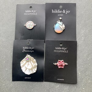 May include: Four pieces of jewellery on black cards. The jewellery includes a connector with iridescent stones, a pendant with a butterfly, a seashell pendant, and a connector with pink stones. The brand name "hildie & jo" is visible.
