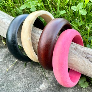 Peut inclure: Quatre bracelets en bois noirs, crème, marron et roses reposent sur un morceau de bois flotté. Les bracelets sont lisses et ont une finition brillante. L'arrière-plan présente de l'herbe verte et une surface en pierre grise.
