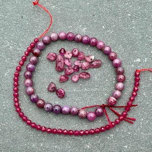 May include: A collection of deep pink and magenta gemstone beads, including a long strand of small faceted round beads, a circular arrangement of larger smooth round beads, and a cluster of irregular, tumbled, and drilled loose gemstone pieces.
