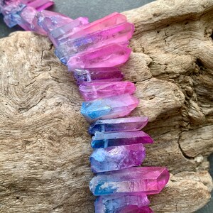 May include: A row of colorful quartz crystals in shades of pink, purple, and blue. The crystals are arranged on a piece of weathered wood, showcasing their natural beauty and vibrant colors.