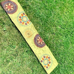 Puede incluir: Un camino de mesa verde con diseños florales bordados en tonos marrones, naranjas y azules. El camino de mesa presenta cinco motivos circulares con patrones intrincados.