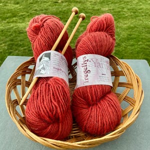 May include: A wicker basket holds two skeins of red yarn labeled "Tregellys Hand Yarns" and two wooden knitting needles. The yarn is a deep, rich red color. The basket sits on a gray surface with a green grassy background.