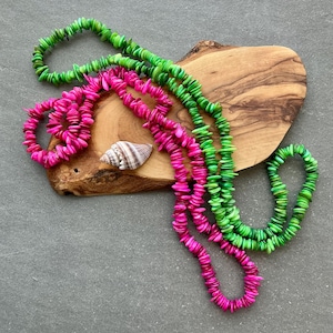 May include: Overhead shot of two necklaces and a bracelet made of small, bright green and fuchsia-colored shell chips. The jewelry is arranged on a wooden cutting board with a seashell. The background is a gray surface.