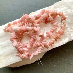 Peut inclure: Un brin de pierres précieuses roses et blanches, probablement du quartz rose ou de l'opale rose, disposé en tas lâche sur une surface blanche.