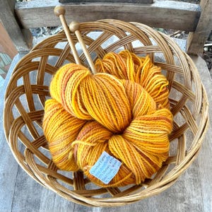 Puede incluir: Una cesta redonda de mimbre contiene un ovillo de lana teñida a mano de color amarillo y naranja y dos agujas de tejer de madera. La etiqueta de la lana dice "Cloverleaf Farms". La lana es una mezcla de 55% Merino y 45% otras fibras.