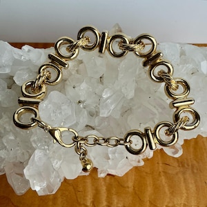 Puede incluir: Pulsera dorada con eslabones circulares y rectangulares entrelazados. La pulsera tiene un cierre de mosquetón y un pequeño dije colgante. El diseño incorpora detalles trenzados en forma de cuerda dentro de los eslabones.