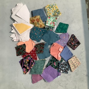 May include: A collection of fabric scraps in various colors and patterns, including floral, geometric, and abstract designs. The scraps are arranged in a random pattern on a light gray surface.