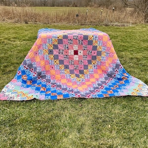 May include: A colorful patchwork quilt with a square pattern, featuring pink, blue, orange, and patterned fabric squares. The quilt is displayed on a grassy surface, showcasing its vibrant design and handcrafted details.