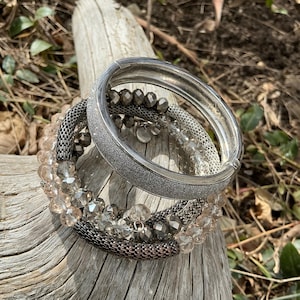 May include: A stack of silver bracelets with various designs. One bracelet is a wide, textured band. Others feature beaded and chain-link styles, with clear and gray beads. The bracelets are resting on a piece of weathered wood.
