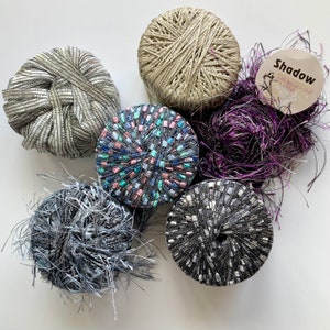 May include: Five spools of yarn in different colors and textures. The spools are arranged in a circle on a white background. The yarn is a mix of silver, purple, black, gray, and pink. The label on one of the spools reads "Shadow, Creative Twist Yarns".