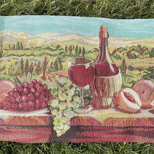 May include: Tapestry depicting a wine scene with a bottle, glasses, grapes, and peaches. The background features a landscape with mountains and trees. The color palette includes shades of red, green, and blue.