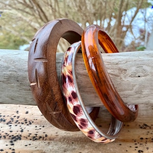 May include: Three bangles of varying styles and materials. One is carved wood, another is a tortoiseshell pattern, and the third is a translucent amber color. The bangles are resting on a piece of driftwood.