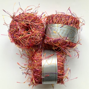 May include: Three skeins of red, orange, yellow, and purple yarn with a silver label that reads "Flutter" and "Made in Italy" with the details "75 yds - 70 m approx, 20 grams ball is standard, 100% Polyester"