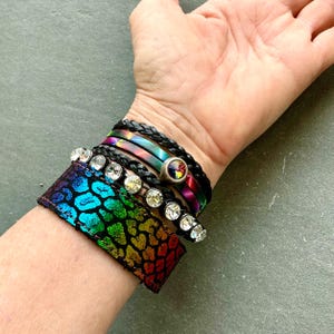 May include: A collection of colorful bracelets. The bracelets include a wide cuff with a rainbow leopard print, a braided black leather wrap, and several thin wraps in iridescent colors. Some bracelets have clear crystal accents.