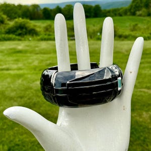以下が含まれることがあります： 幾何学模様の黒とシルバーのブレスレット。ブレスレットは長方形で、ライトブルーのアクセントが付いています。ブレスレットは、緑の背景に白い手で表示されています。