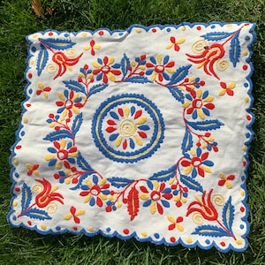 May include: White square embroidered textile with a scalloped edge. The embroidery features a central circular design and floral patterns in red, yellow, and blue. The textile is placed on a green grassy surface.