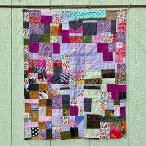 May include: A patchwork quilt with a variety of fabrics in shades of purple, pink, brown, and green. The quilt is made of squares and rectangles of different sizes and patterns, including floral, polka dots, and geometric designs. The quilt is hanging on a light green wooden wall.