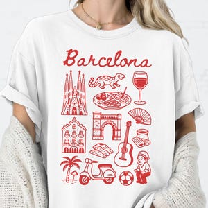 May include: White t-shirt with a red Barcelona graphic. The design includes illustrations of landmarks, food, and cultural symbols. The word "Barcelona" is written in a script font above the illustrations.