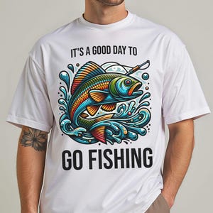 May include: White t-shirt with a colorful graphic of a fish jumping out of water, with the text "IT'S A GOOD DAY TO GO FISHING". The fish has blue, orange, and green scales. The shirt is a casual style.