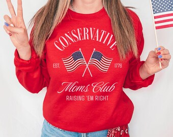 May include: A red crewneck sweatshirt featuring white text that reads CONSERVATIVE, Moms Club, and RAISING 'EM RIGHT, with the text EST. 1776 and a graphic of two crossed American flags printed on the front center of the garment.