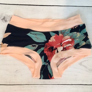 May include: Floral print underwear with a peach colored waistband. The underwear has a dark blue background with a floral pattern in shades of red, green, and white.