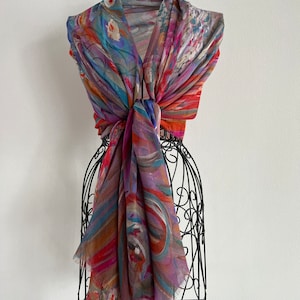 A colorful scarf with abstract patterns in shades of red, orange, blue, and purple. This artful elegant scarf is a silk modal all season blend and is draped over a black metal mannequin.