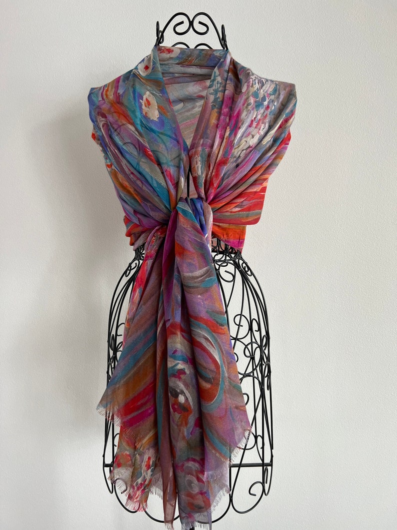 A colorful scarf with abstract patterns in shades of red, orange, blue, and purple. This artful elegant scarf is a silk modal all season blend and is draped over a black metal mannequin.