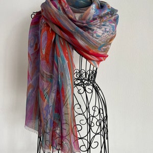 A colorful scarf with abstract patterns in shades of red, orange, blue, and purple. This artful elegant scarf is a silk modal all season blend and is draped over a black metal mannequin.