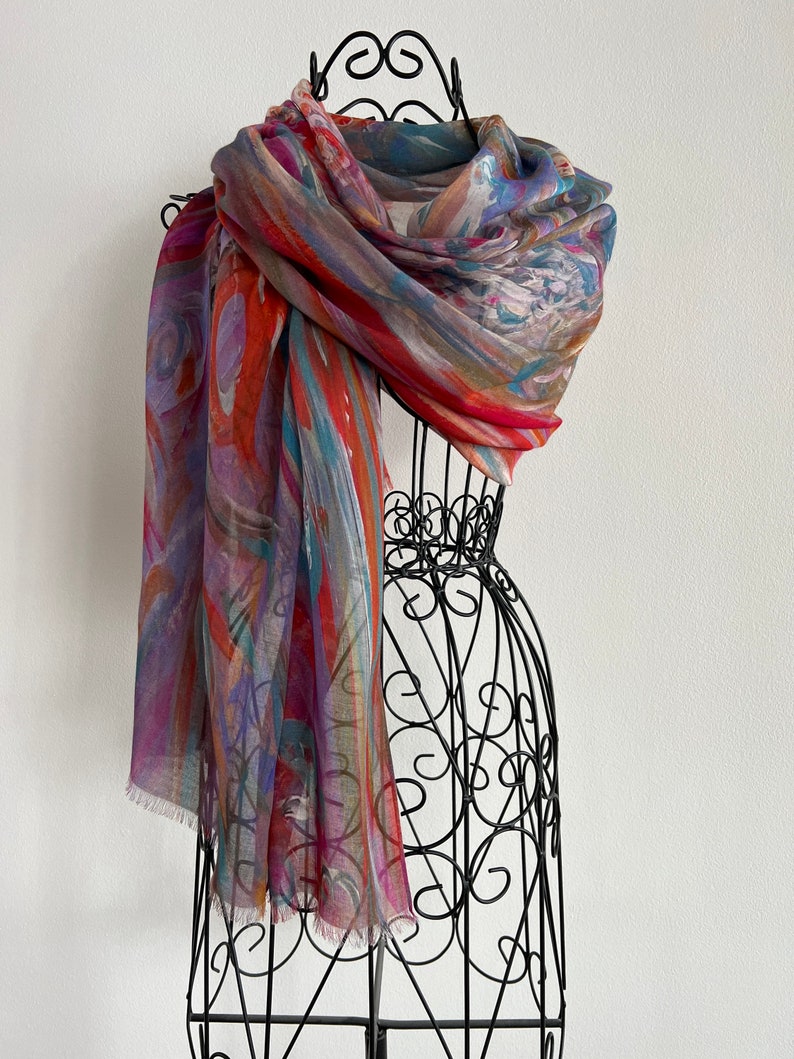 A colorful scarf with abstract patterns in shades of red, orange, blue, and purple. This artful elegant scarf is a silk modal all season blend and is draped over a black metal mannequin.