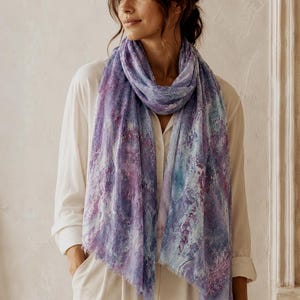 May include: A lightweight scarf featuring an abstract marble-like pattern in shades of purple, lavender, and light blue. The scarf has a soft, semi-sheer texture with delicate frayed edges along the bottom hem, draped loosely around the neck.