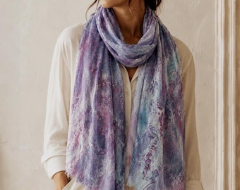 May include: A lightweight scarf featuring an abstract marble-like pattern in shades of purple, lavender, and light blue. The scarf has a soft, semi-sheer texture with delicate frayed edges along the bottom hem, draped loosely around the neck.