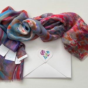 A colorful silk modal scarf with a marbled pattern in shades of blue, pink, red, and orange. The scarf is folded and displayed on a white gift envelope with a heart-shaped logo featuring a stylized flower design. The sticker has the text 'The Big Soul