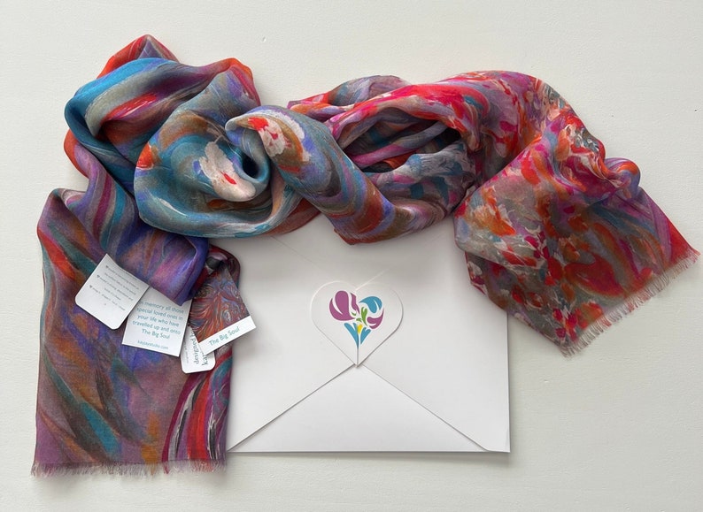 A colorful silk modal scarf with a marbled pattern in shades of blue, pink, red, and orange. The scarf is folded and displayed on a white gift envelope with a heart-shaped logo featuring a stylized flower design. The sticker has the text 'The Big Soul