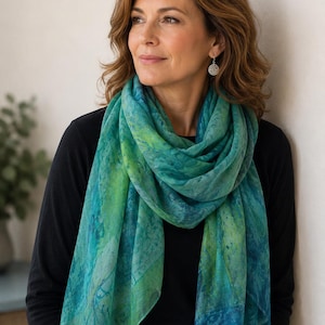 May include: A lightweight rectangular scarf featuring a marbled pattern in shades of teal, emerald green, and lime green. The scarf has a soft, semi-sheer texture with delicate frayed edges along the bottom hem, worn over a solid black long-sleeve top.