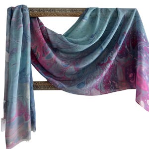 May include: A rectangular scarf featuring a marbled pattern with swirling shades of blue, teal, and magenta. The fabric is draped over a rectangular gold-toned frame, displaying a fluid, abstract design with visible paint-like textures throughout.