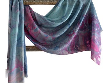 May include: A rectangular scarf featuring a marbled pattern with swirling shades of blue, teal, and magenta. The fabric is draped over a rectangular gold-toned frame, displaying a fluid, abstract design with visible paint-like textures throughout.