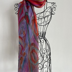 A colorful scarf with abstract patterns in shades of red, orange, blue, and purple. This artful elegant scarf is a silk modal all season blend and is draped over a black metal mannequin.