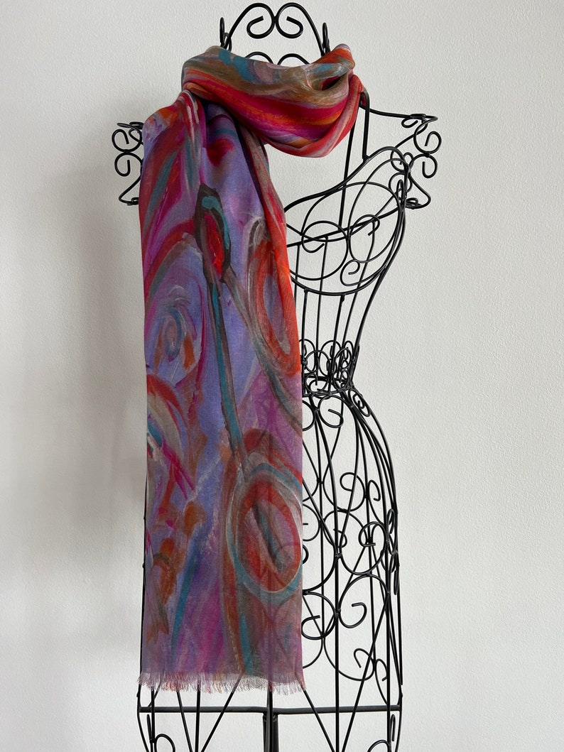 A colorful scarf with abstract patterns in shades of red, orange, blue, and purple. This artful elegant scarf is a silk modal all season blend and is draped over a black metal mannequin.