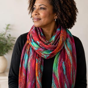 May include: A rectangular scarf featuring a vibrant, abstract pattern with bold streaks of deep red, magenta, bright turquoise, and hints of orange. The fabric has a soft, lightweight appearance with a fringed edge, draped loosely around the neck.