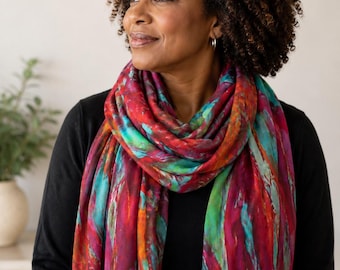 Puede incluir: Una bufanda rectangular con un patrón abstracto vibrante que presenta rayas audaces en rojo intenso, magenta, turquesa brillante y toques de naranja. La tela tiene una apariencia suave y ligera con un borde deshilachado alrededor del cuello.
